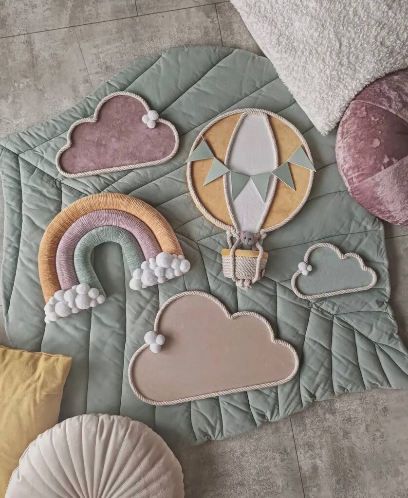 Zsályazöld játszószőnyegen mustársárga hőlégballon dísz, mellette pamutzsinóros szivárvány és felhők, felülnézetből